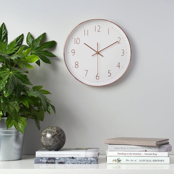 A white wall clock with copper accents displays time on a grey wall, flanked by a potted plant and stacked books on a shelf.