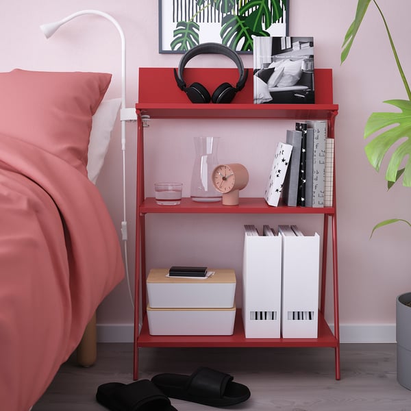 Bedside shelf: KRÖNIKÖR red, holds books, clock, headphones, small plants, glass, box. Tall, slim shape, bright colour. Next to bed, smaller than bed.