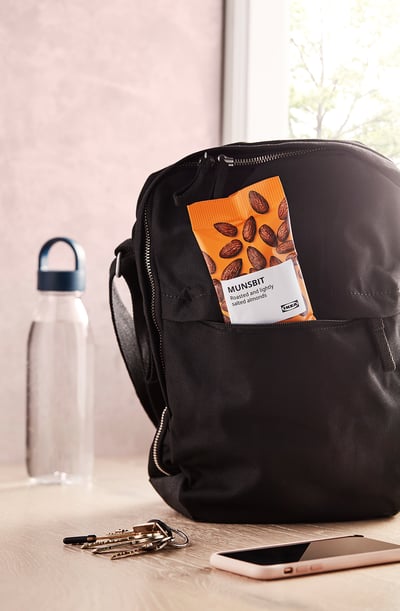 Backpack with orange snack pack, keys, and phone on light table. A window shows trees outside.