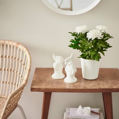 A wooden table holds white figures and a potted plant with white flowers. A wicker chair sits beside it with a mirror above.