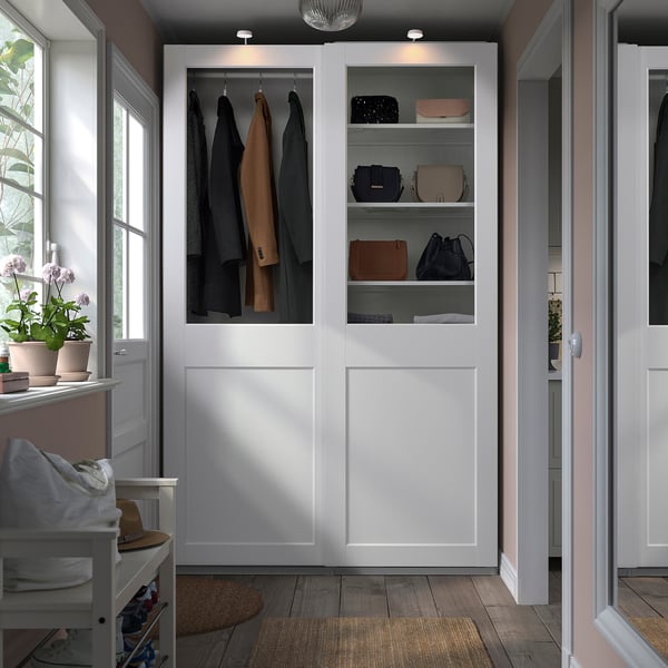 A modern white PAX wardrobe, tall with glass doors. It holds coats and handbags neatly. A chair sits nearby, small in comparison.