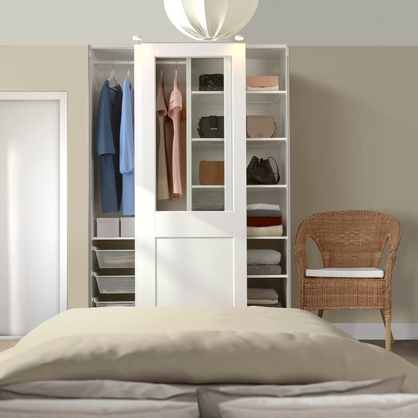 White PAX wardrobe with glass doors, beside bed and chair, holding clothes and bags.