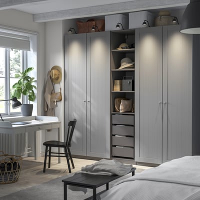 Modern bedroom with grey PAX wardrobe, integrated lighting, and storage. Beside bed: lamp, plant, hats, bags.