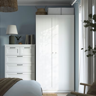 Modern bedroom featuring PAX wardrobe, dresser, and bed. Wardrobe dominates room, holding lamp, books, and decor.