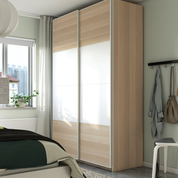 Modern bedroom with tall, light wood PAX closet against green wall, next to window. Includes bed, stool, and hooks with clothes.