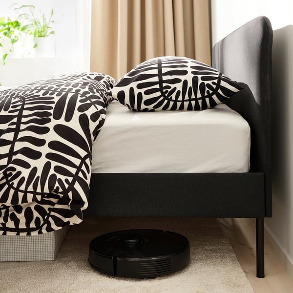 Bedroom scene with modern bed, black robot vacuum, cushions, and curtains. Vacuum under bed highlights easy cleaning.