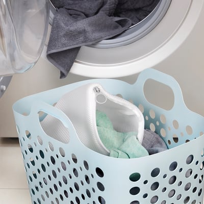 Light blue perforated laundry basket with white mesh bag inside, sitting near open washing machine filled with clothes.