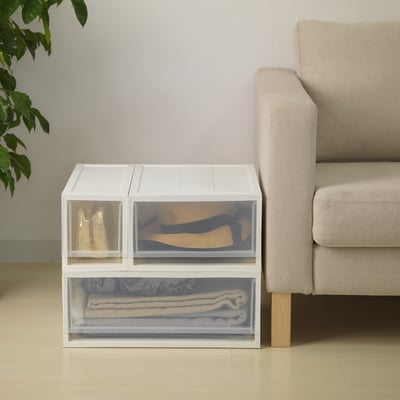 Clear storage unit stacked with folded items beside neutral sofa.