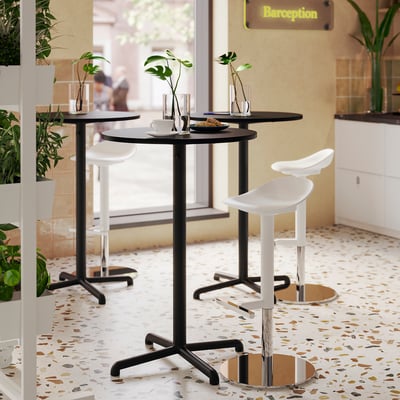A modern cafe with black tables and white stools, surrounded by plants. Yellow sign in background.