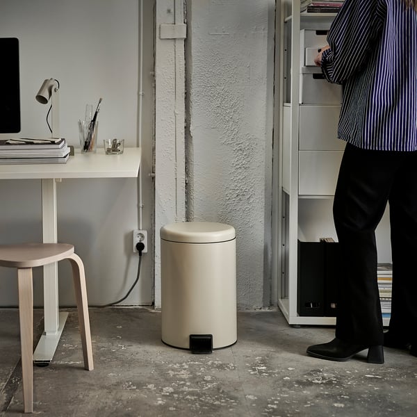 Person standing near white rubbish can with pedal, a desk with a computer and stool nearby.