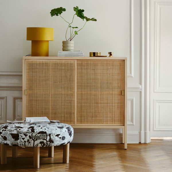 STOCKHOLM 2025 lamp in sun-yellow linen on wooden cabinet, contrasting shade, with greenery, books, and patterned ottoman.