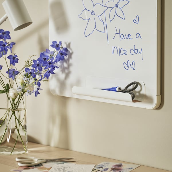 Small white VEMUND pen holder on magnetic board, holding blue pen, scissors. Next to small vase with purple flowers and lamp.