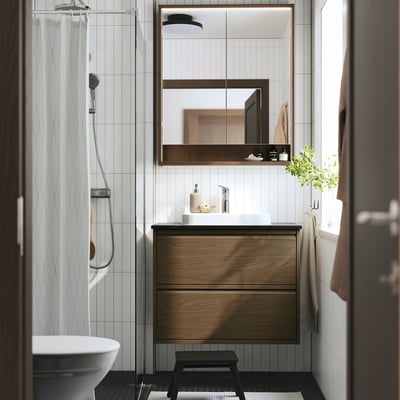 Modern bathroom with ÄNGSJÖN oak wash-stand, white marble basin, chrome shower, dark cabinet, and stool. Soft light, calm space.