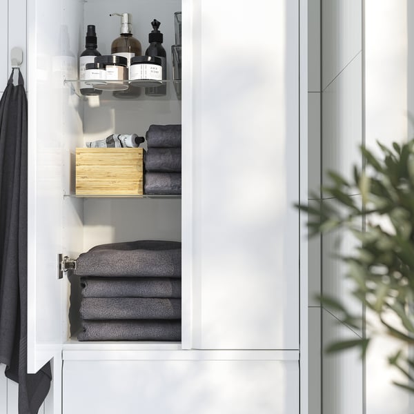 Open white bathroom cabinet displaying grey towels, a wooden box, and bottles on shelves, with a plant nearby.