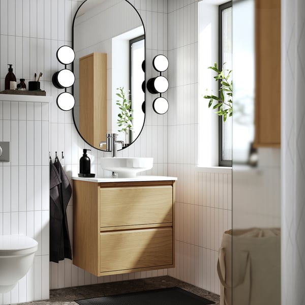 Modern bathroom with light wood vanity, round mirror, vessel sink, and black accents. Spacious, organised drawers enhance minimalist design.