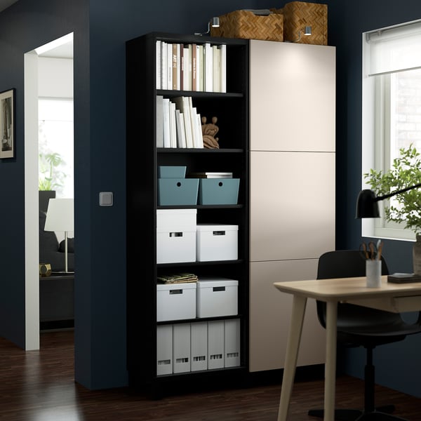A tall black BESTÅ unit with white doors in a modern room, holding storage boxes and books, next to a desk and chair.
