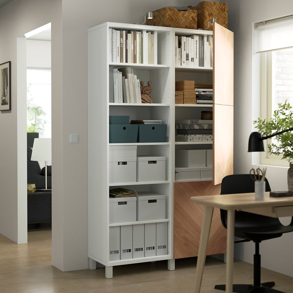 A modern home office a BESTÅ bookcase with adjustable shelves holding books and storage boxes, adjacent to a desk with a chair and plants.