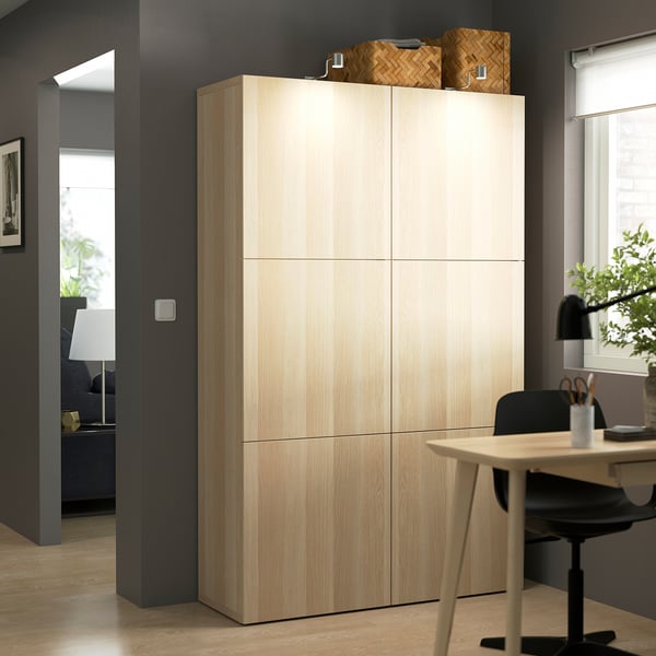 Large wooden BESTÅ cabinet with six doors, taller than desk and lamp, two baskets on top.