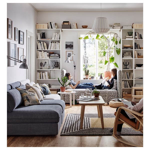 Modern living room with grey sectional, wooden tables, striped rug, bookcases, hanging lamp, white walls, and windows with greenery views.