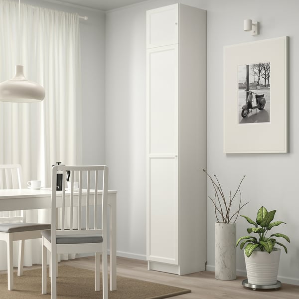 Minimalist dining area with white BILLY bookcase, potted plant, framed scooter photo, and white chairs around a table.