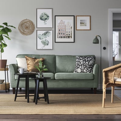 Modern living room with EKHOLMA sofa, green, plush, 3-seater. Wall art, floor lamp, wicker chair, and rug. Clean, minimalist design.