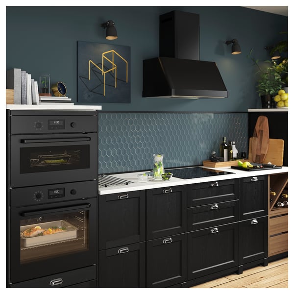 Modern kitchen with dual ovens, black cabinets, blue backsplash, and a black range hood. Decorated with plants and artwork.