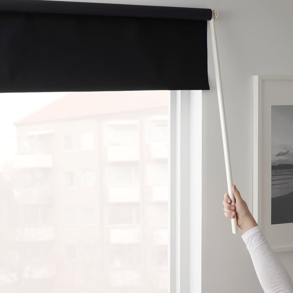 A person raises a wand to adjust a black roller blind, blocking sunlight from a wide window.
