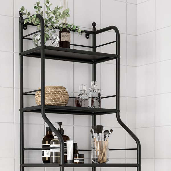 Black metal bathroom shelf unit with three tiers storing towels, bottles, vases, and makeup brushes against white tiled wall.