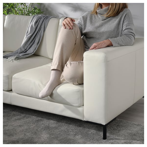 A person sits on a modern white leather couch, wearing a grey sweater and beige pants, with a grey throw blanket on the seat next to them.