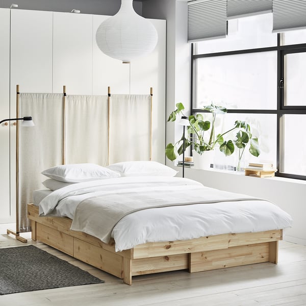 Minimalist bedroom with white GLAMBERGET extendable bed, light wood frame, beige curtains, white linens, large window with plants, black rug.