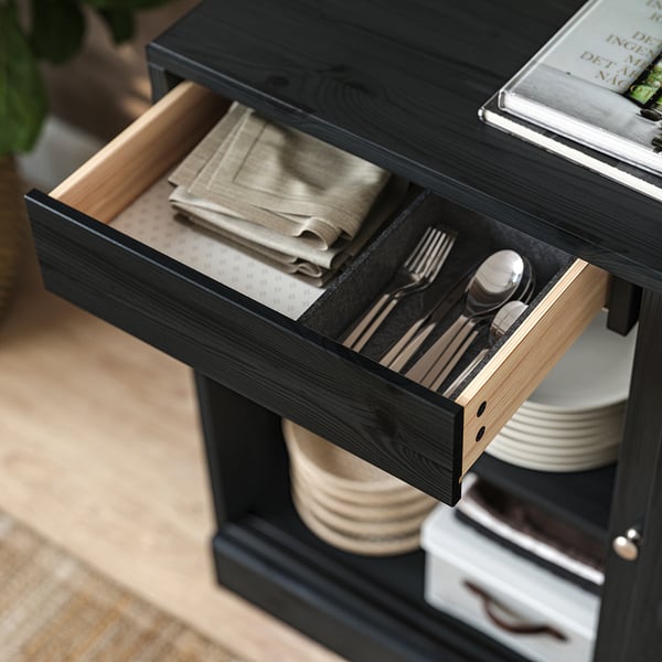 Black HAVSTA sideboard drawer open, showing cutlery and beige napkins, made of solid pine.