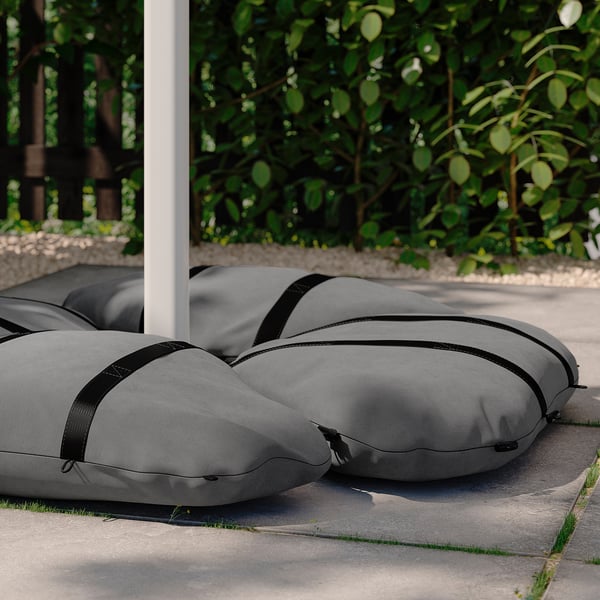 Grey hanging parasol covers on paving, surrounded by greenery. Cylindrical with black straps for easy carrying.