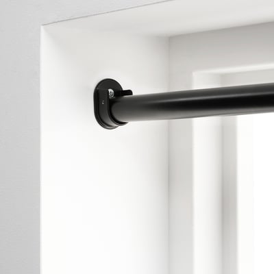 Black curtain rod bracket mounted on white wall, part of HUGAD system for hanging curtains between two opposite walls.