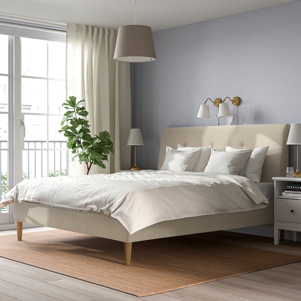 Modern beige upholstered IDANÄS bed with sloping headboard, white linens, nightstands, and large window. Cosy, minimalist design.