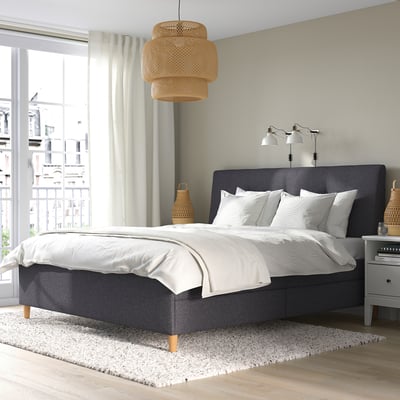 IDANÄS bed in dark grey with upholstered headboard & wooden legs in bright room with large window.