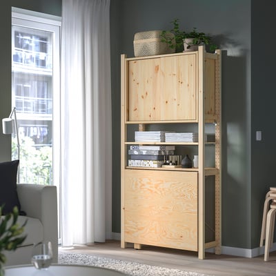 IVAR pine storage unit stands tall with a simple design, holding stacks of books and a plant, fitting neatly near a large window against green walls.