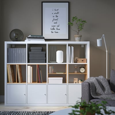 Modern living room with KALLAX shelf filled with books, decor, and plants. Grey sofa with blanket, cosy and organised.