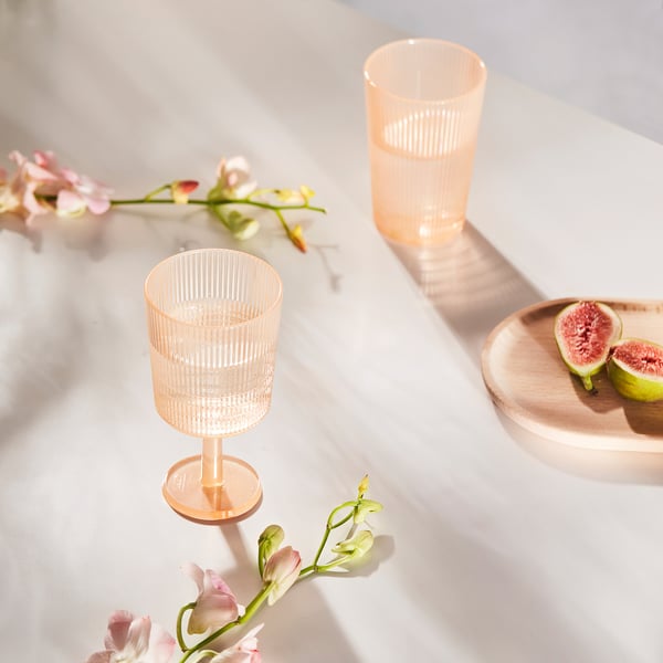 Two pink plastic wine glasses, one strawberry on a wooden plate, surrounded by flowers on a marble table.