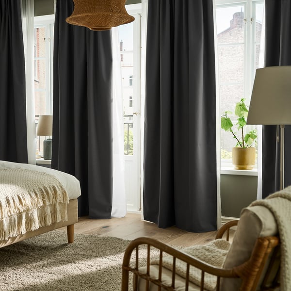 Modern bedroom with dark curtains, wooden bed, wicker chair, city view from tall window, green plant.