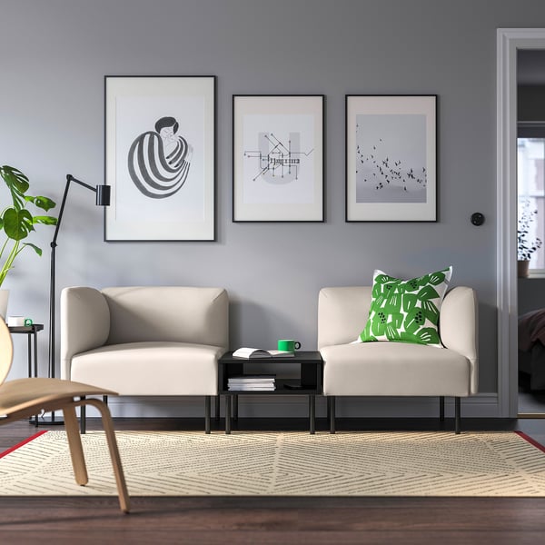 Modern living room with neutral sofas, green pillows, black coffee table, abstract art, grey walls, wooden floor, rug, and tall lamp with plant.