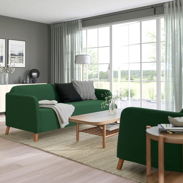 Modern living room with green sofa, pillows, coffee table, armchair, side table with books, and rug on light wood floor.
