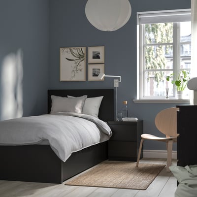 Modern minimalist bedroom with blue walls, black MALM bed, grey bedding, wooden chair, and window with plants.