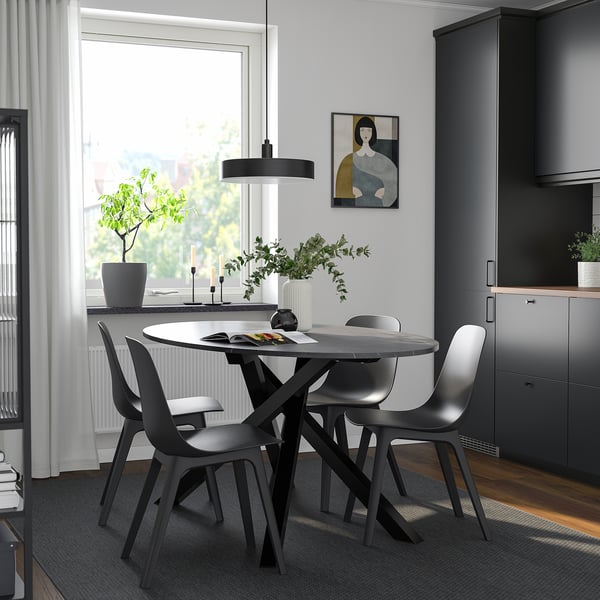 Modern dining setup with round table, four black chairs, pendant light, grey rug, and large window with natural light and artwork.