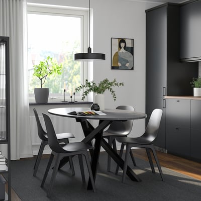 Modern dining setup with round table, four black chairs, pendant light, grey rug, and large window with natural light and artwork.