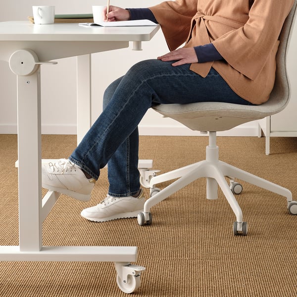 Person sitting at adjustable table with footrest, taking notes. White trainers, jeans, and sweater visible.