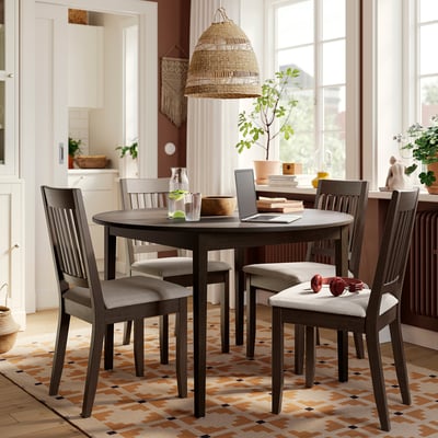 Modern farmhouse dining set with dark grey round table and 4 cushioned chairs, placed on a multicoloured rug under a large window.