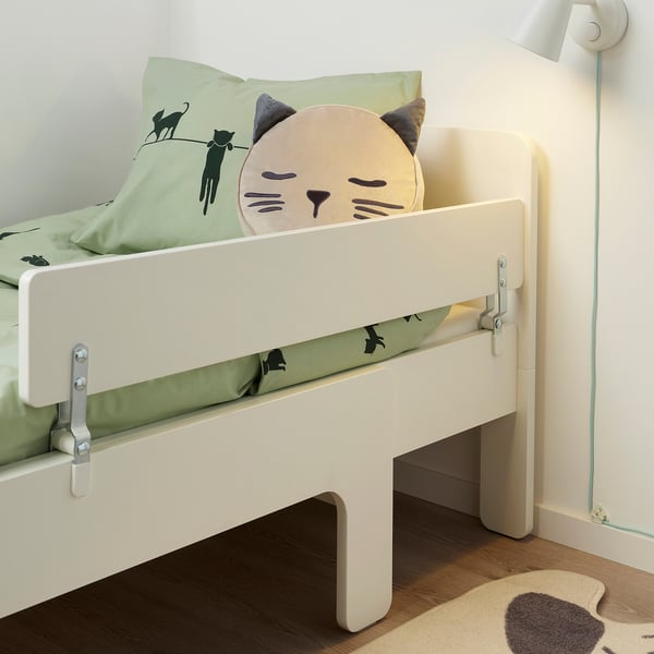 A beige childrens bed with a side guardrail, showcasing a green pillow with cat illustrations and a cat-themed cushion.