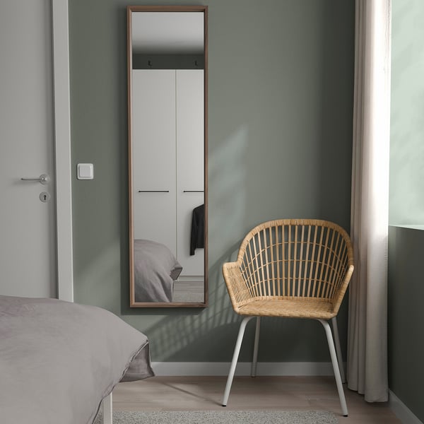 Minimalist bedroom with NISSEDAL mirror, wicker chair, and wardrobe. Mirror leans on green wall, reflecting bed and wardrobe.