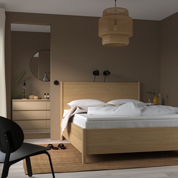 Cosy bedroom with wooden NORDBERGET bed, orthopaedic mattress, white sheets, pendant light, dresser with mirror, black chair, and brown rug.
