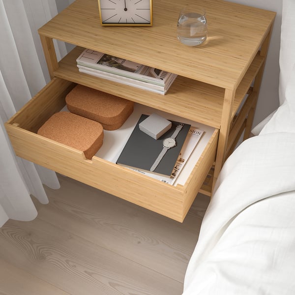 Bedside table with open drawer showing magazines and cork blocks. Clock and glass on top, white bed linens nearby.